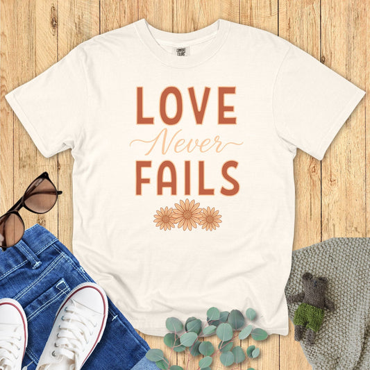 Flatlay of a Comfort Colors 1717 ivory “Love Never Fails – 1 Corinthians 13:8” t-shirt with rust orange text and daisy flower details. A cozy women’s scripture tee — soft modern Christian shirt and lovely faith-based apparel for women, perfect for Christian mom gift ideas.