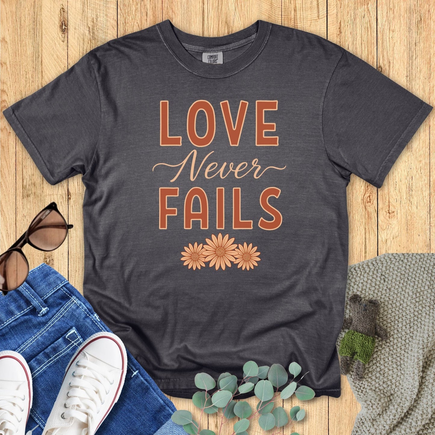Flatlay of a Comfort Colors 1717 pepper “Love Never Fails – 1 Corinthians 13:8” t-shirt with soft orange lettering and floral accents. A minimalist women’s scripture tee — modern Christian shirt and elegant faith-based apparel for women, perfect Christian mom gift idea.