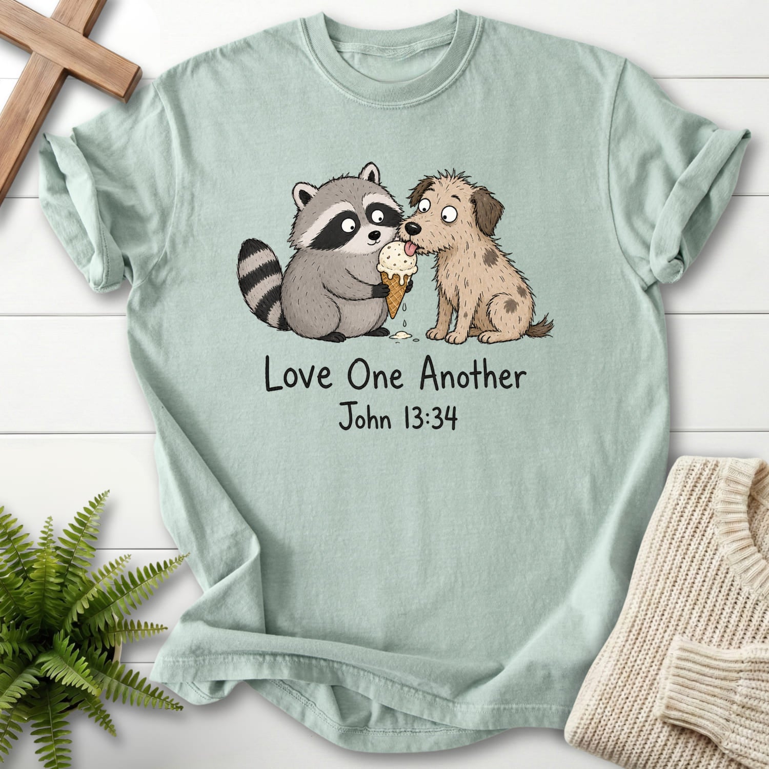 Flat lay of a Bay women's Christian tee featuring "Love One Another Raccoon Puppy T-Shirt" John 13:34.