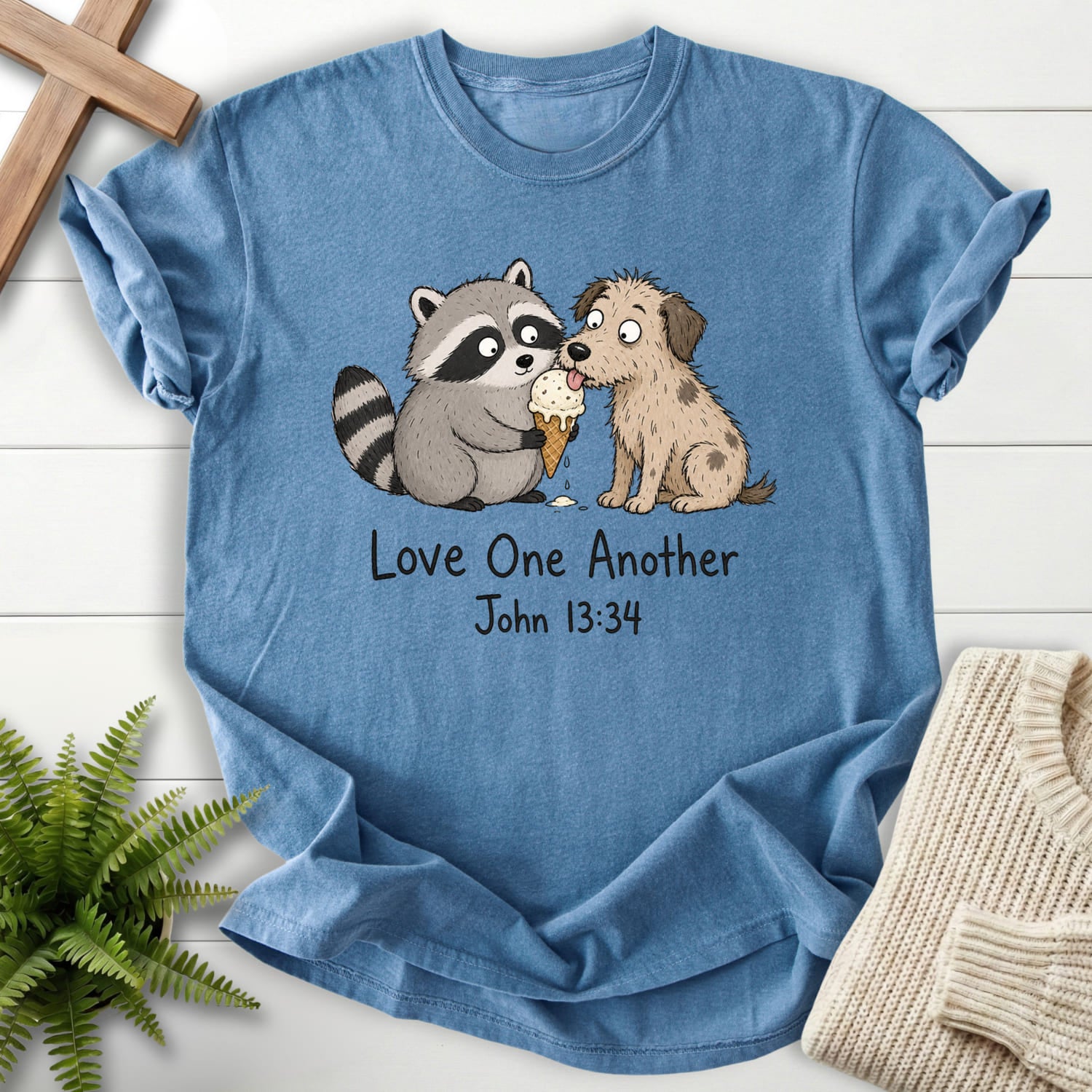 Flatlay of a Blue Jean faith-inspired shirt with "Love One Another Raccoon Puppy T-Shirt" John 13:34.