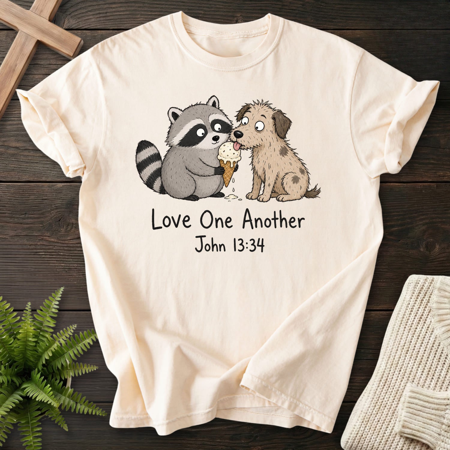 Flatlay of an Ivory faith-based tee with "Love One Another Raccoon Puppy T-Shirt" John 13:34.