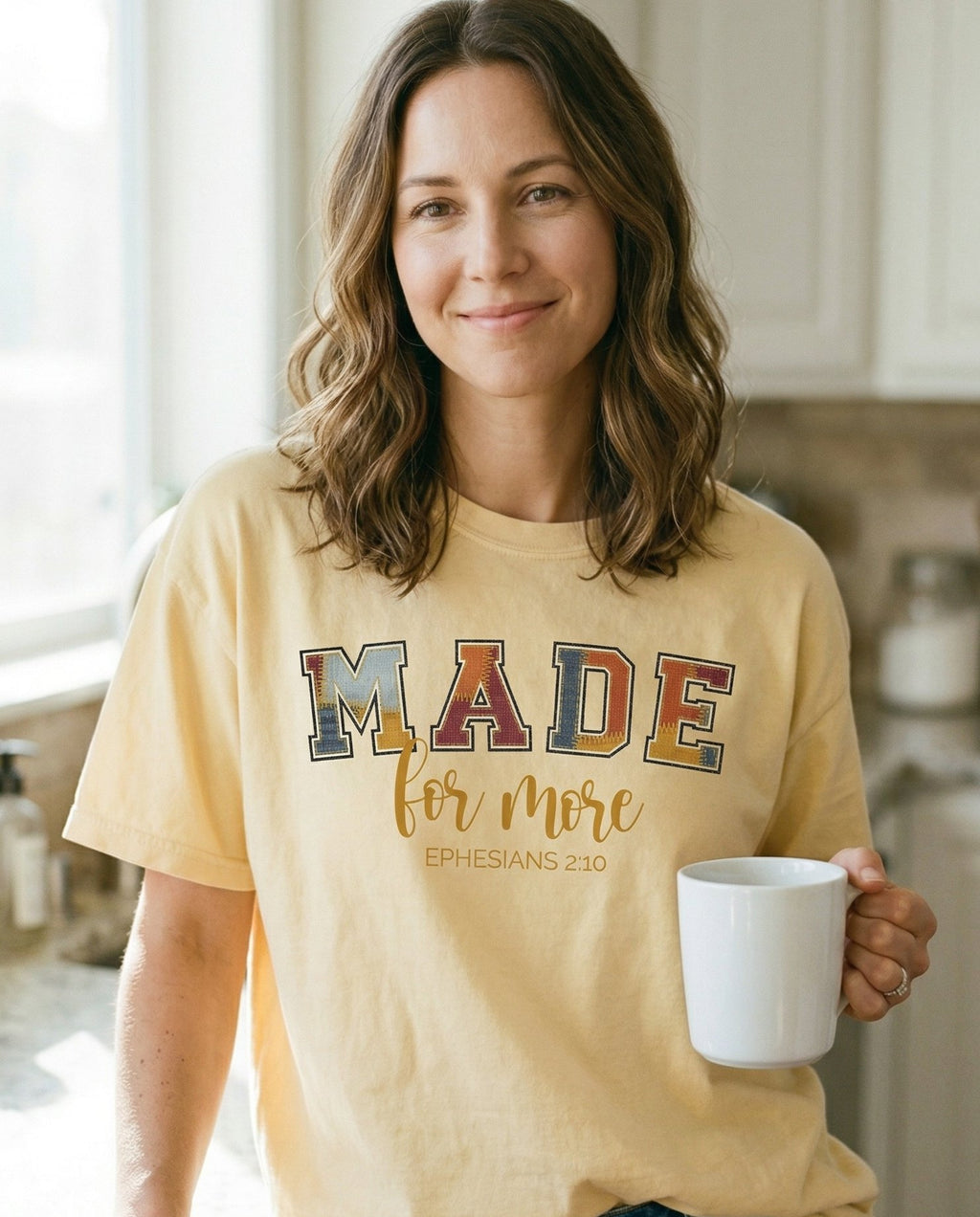 Made for More – Banana women's Christian tee, lifestyle photo in morning kitchen setting – Varsity-style patchwork lettering spelling 'MADE' in multicolored text – Ephesians 2:10 – She Wears Faith