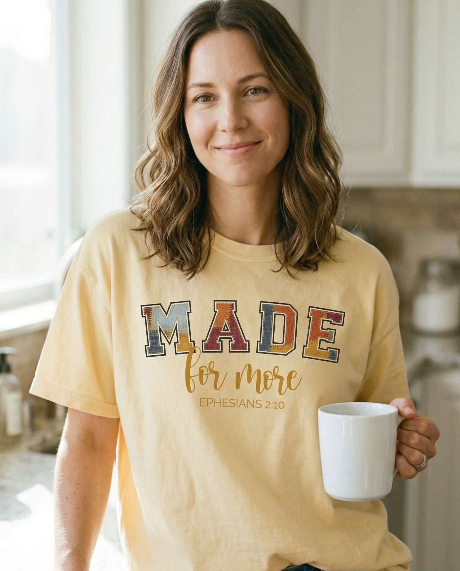 Made for More – Banana women's Christian tee, lifestyle photo in morning kitchen setting – Varsity-style patchwork lettering spelling 'MADE' in multicolored text – Ephesians 2:10 – She Wears Faith