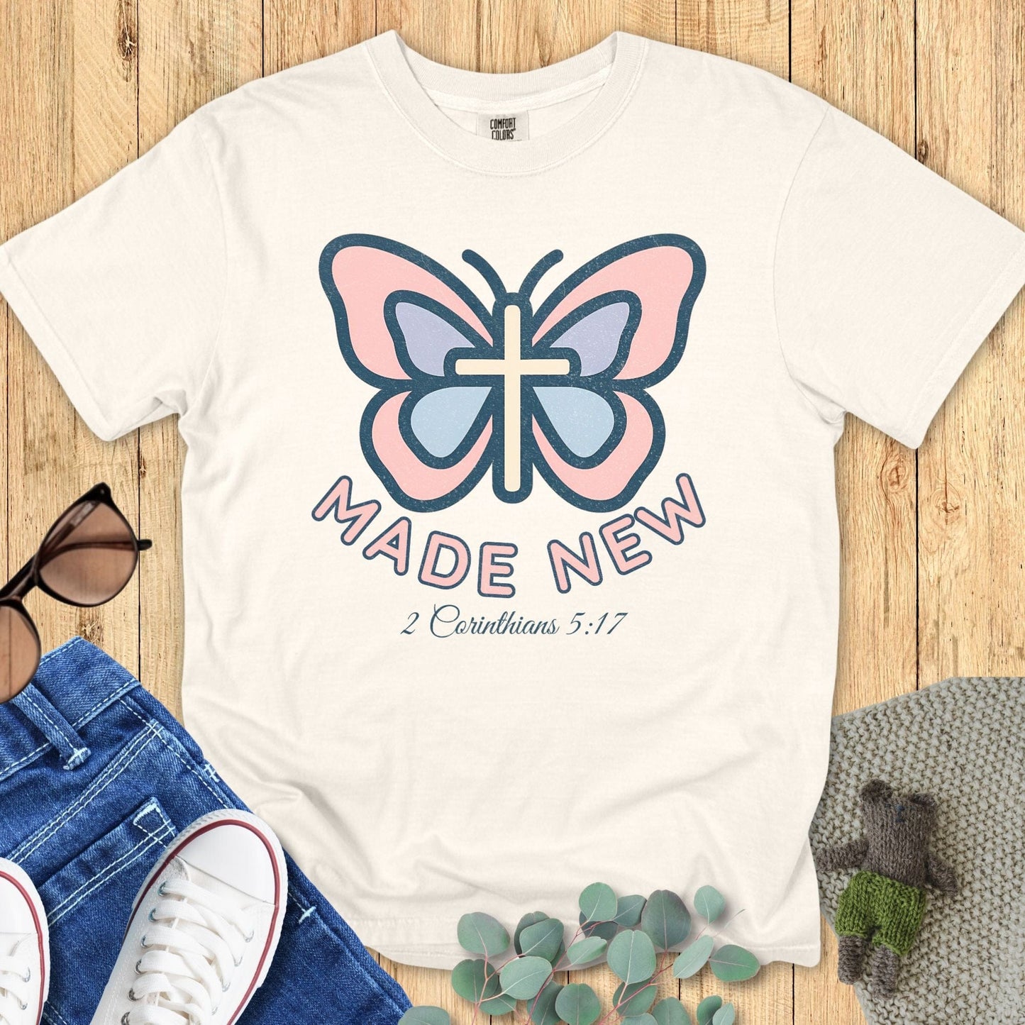 Flat lay of a Comfort Colors 1717 ivory Christian women’s t-shirt with a butterfly and cross graphic, text “Made New – 2 Corinthians 5:17”, styled with jeans and sneakers, boutique Christian apparel.