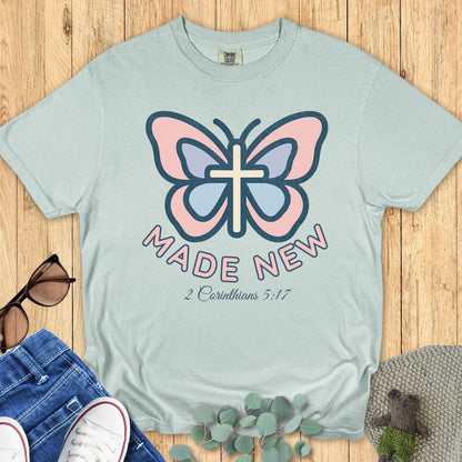 Flat lay of a Comfort Colors 1717 bay blue Christian women’s t-shirt with a butterfly and cross graphic, text “Made New – 2 Corinthians 5:17”, boutique Christian apparel design.
