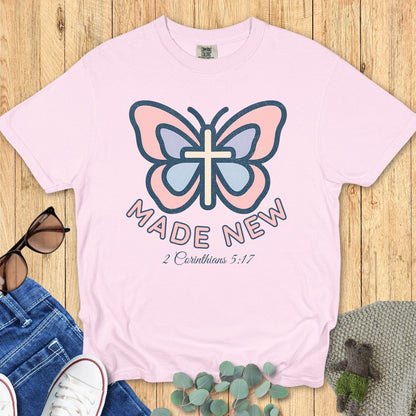 Flat lay of a Comfort Colors 1717 blossom pink Christian women’s t-shirt with butterfly and cross graphic, text “Made New – 2 Corinthians 5:17”, feminine Christian apparel style.