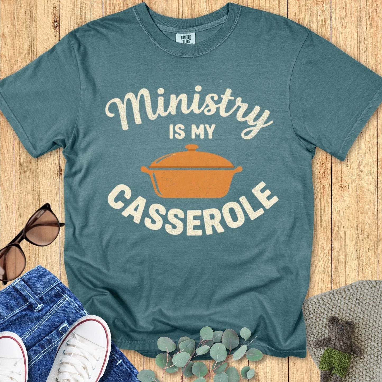 Teal t-shirt with 'Ministry is my Casserole' graphic on a wooden surface with sunglasses and jeans.
