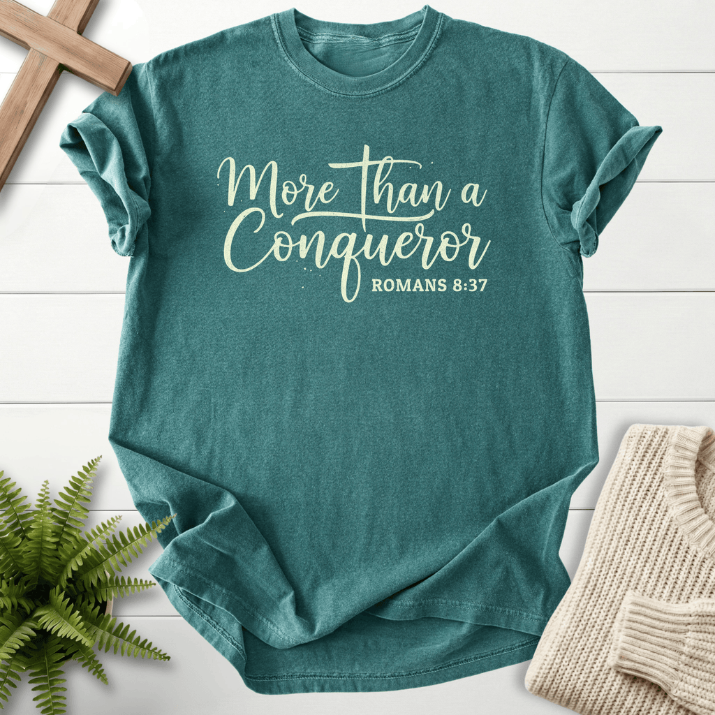 Blue Spruce women's Bible verse tee with cursive 'More Than a Conqueror' lettering, inspired by Romans 8:37.