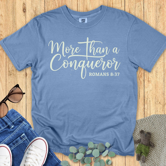 Comfort Colors 1717 blue jean “More Than a Conqueror – Romans 8:37” t-shirt with creamy white text in cursive script. A stylish women’s scripture tee — empowering faith-based apparel for women and encouraging modern Christian shirt, ideal for Christian mom gift ideas.