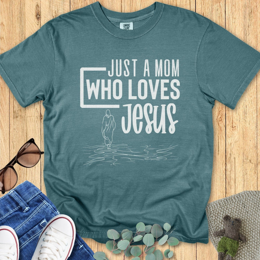 Flatlay of a Comfort Colors 1717 blue spruce “Just a Mom Who Loves Jesus” t-shirt with subtle line art of Jesus walking on water. A soft women’s scripture tee — faith-based apparel for women and modern Christian shirt, thoughtful Christian mom gift idea.
