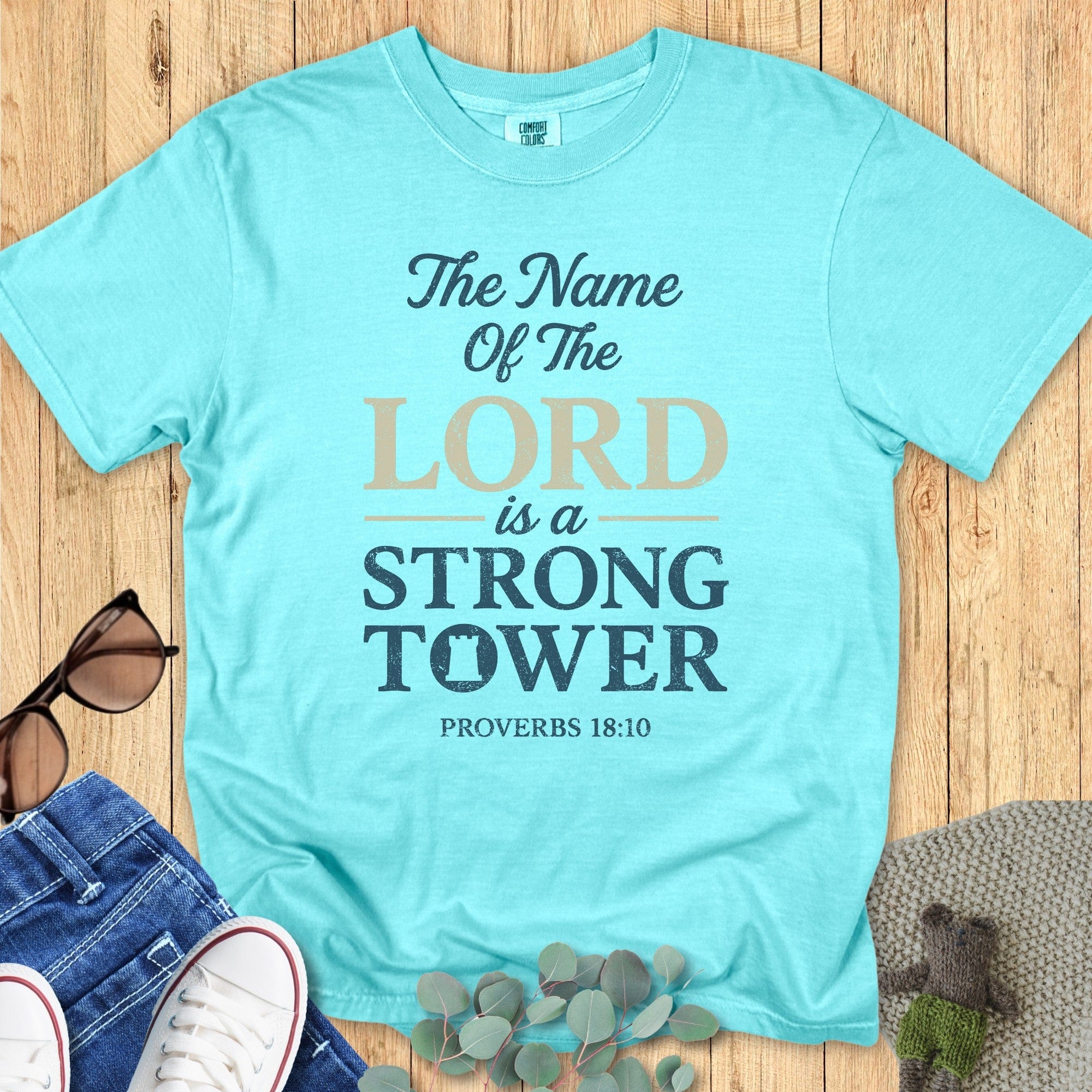 Woman standing near a peaceful lake wearing a Comfort Colors 1717 aqua “The Name of the Lord is a Strong Tower – Proverbs 18:10” t-shirt. A modern Christian shirt from faith-based apparel for women collection.