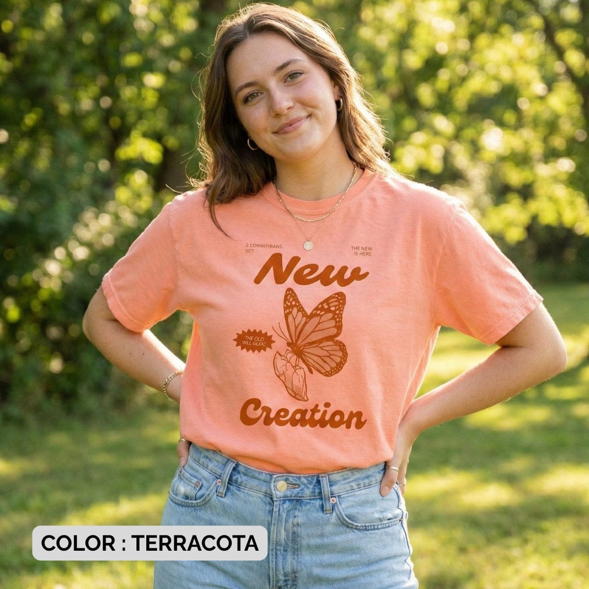Christian woman wearing a terracotta Comfort Colors 1717 “New Creation” tshirt featuring a butterfly and 2 Corinthians 5:17 design, standing outdoors in soft natural light. Stylish US faith wear aesthetic with a warm, encouraging message about new life in Christ.