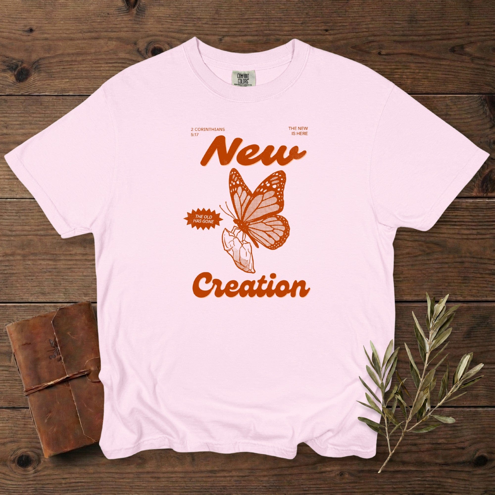 Christian women’s tshirt in soft blossom pink Comfort Colors 1717 showing a retro “New Creation” butterfly illustration inspired by 2 Corinthians 5:17. Faith-based graphic with warm orange ink, styled flat lay with vintage journal and olive leaves, modern US faith wear.