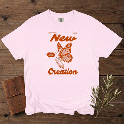 Christian women’s tshirt in soft blossom pink Comfort Colors 1717 showing a retro “New Creation” butterfly illustration inspired by 2 Corinthians 5:17. Faith-based graphic with warm orange ink, styled flat lay with vintage journal and olive leaves, modern US faith wear.
