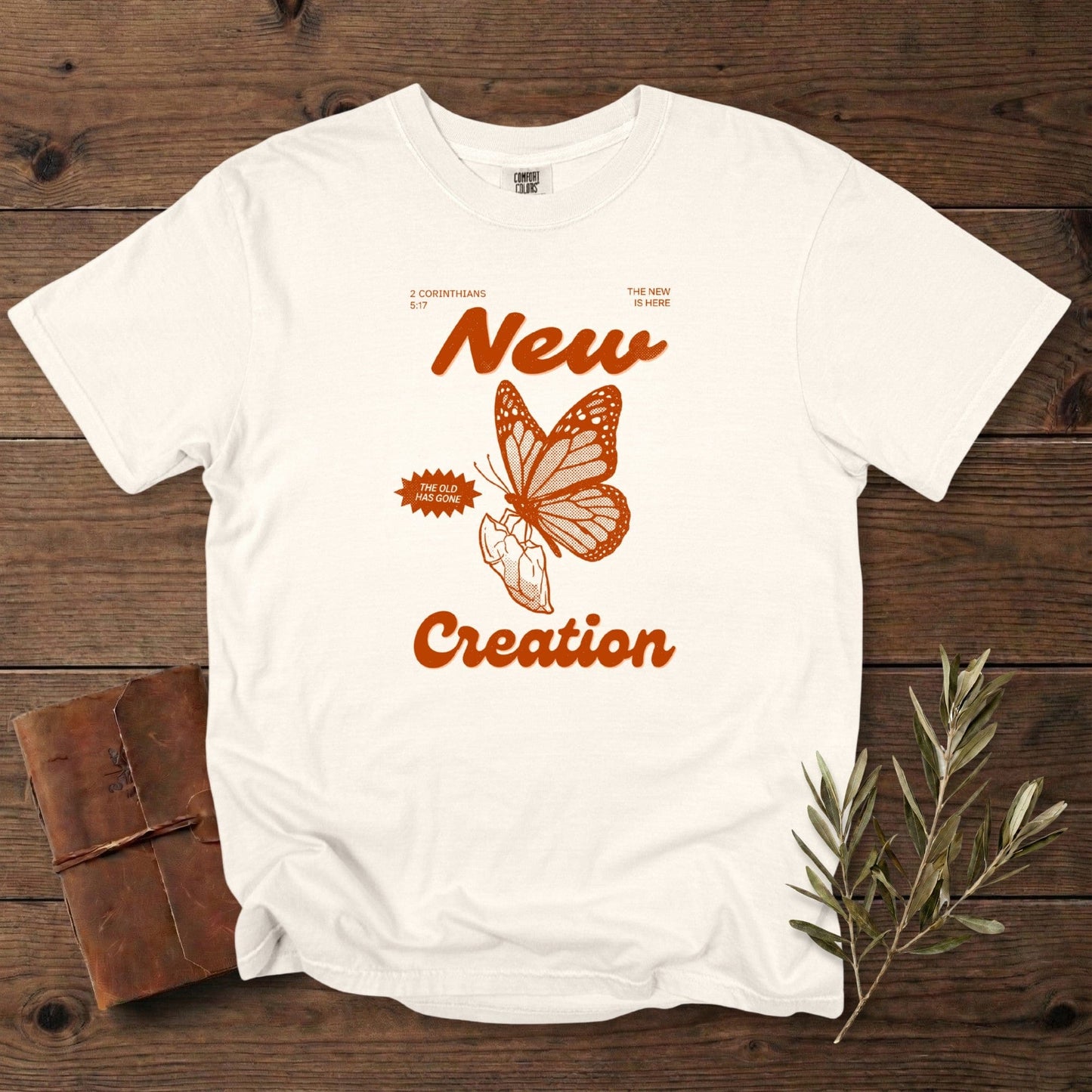 Ivory Christian women tee on Comfort Colors 1717 with “New Creation” text and a bold butterfly-and-hand illustration referencing 2 Corinthians 5:17. Minimal vintage faith design, perfect for US faith wear, displayed on a rustic wood backdrop with olive branches.