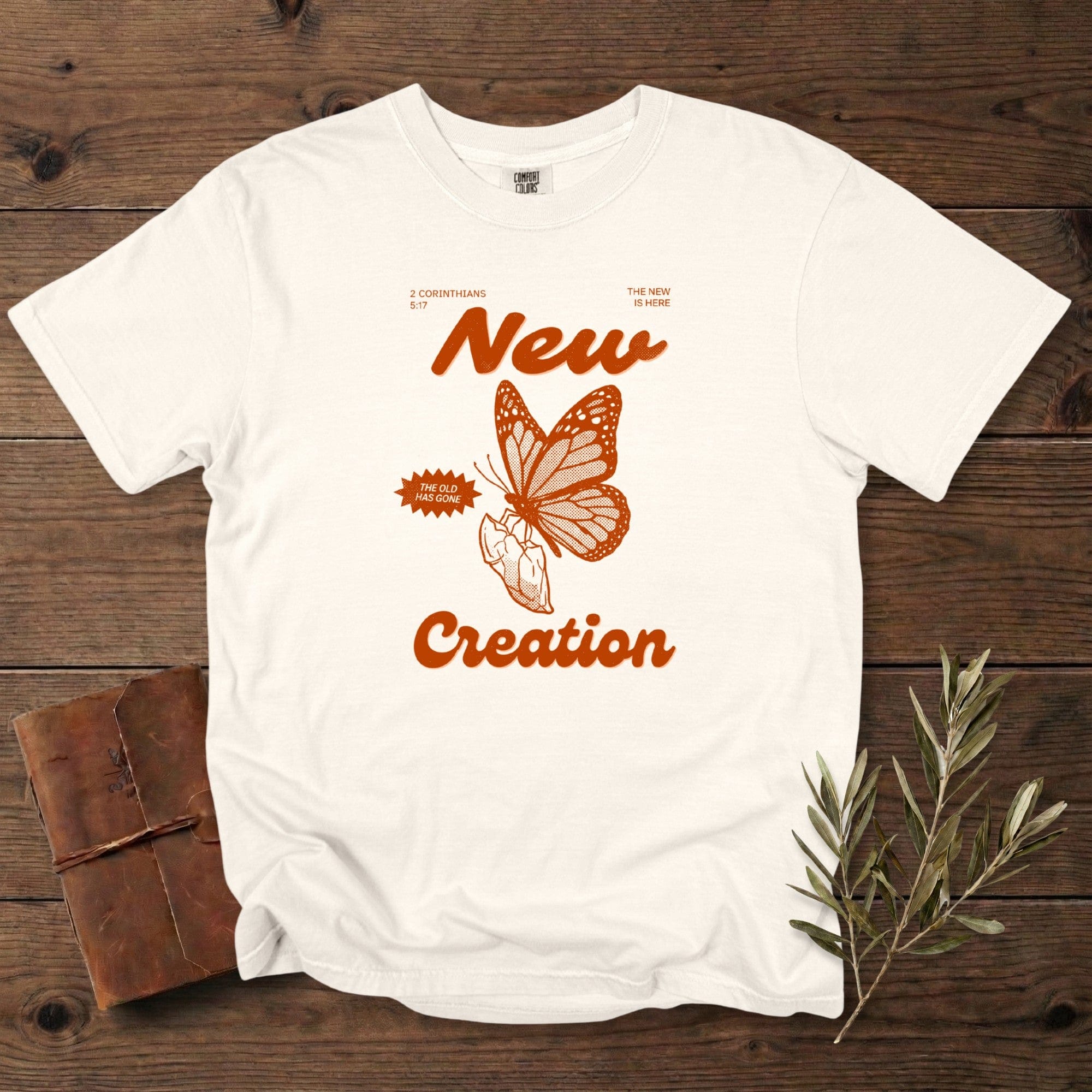 Ivory Christian women tee on Comfort Colors 1717 with “New Creation” text and a bold butterfly-and-hand illustration referencing 2 Corinthians 5:17. Minimal vintage faith design, perfect for US faith wear, displayed on a rustic wood backdrop with olive branches.