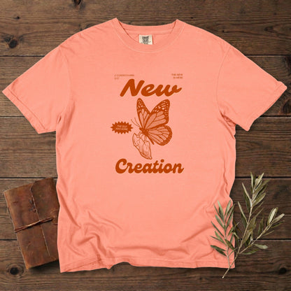 Christian women’s tee in vintage terracotta Comfort Colors 1717 featuring a “New Creation” design inspired by 2 Corinthians 5:17, with a hand-drawn butterfly emerging from a hand. Retro faith graphic, US faith wear aesthetic, photographed on a rustic wood background with olive branches.