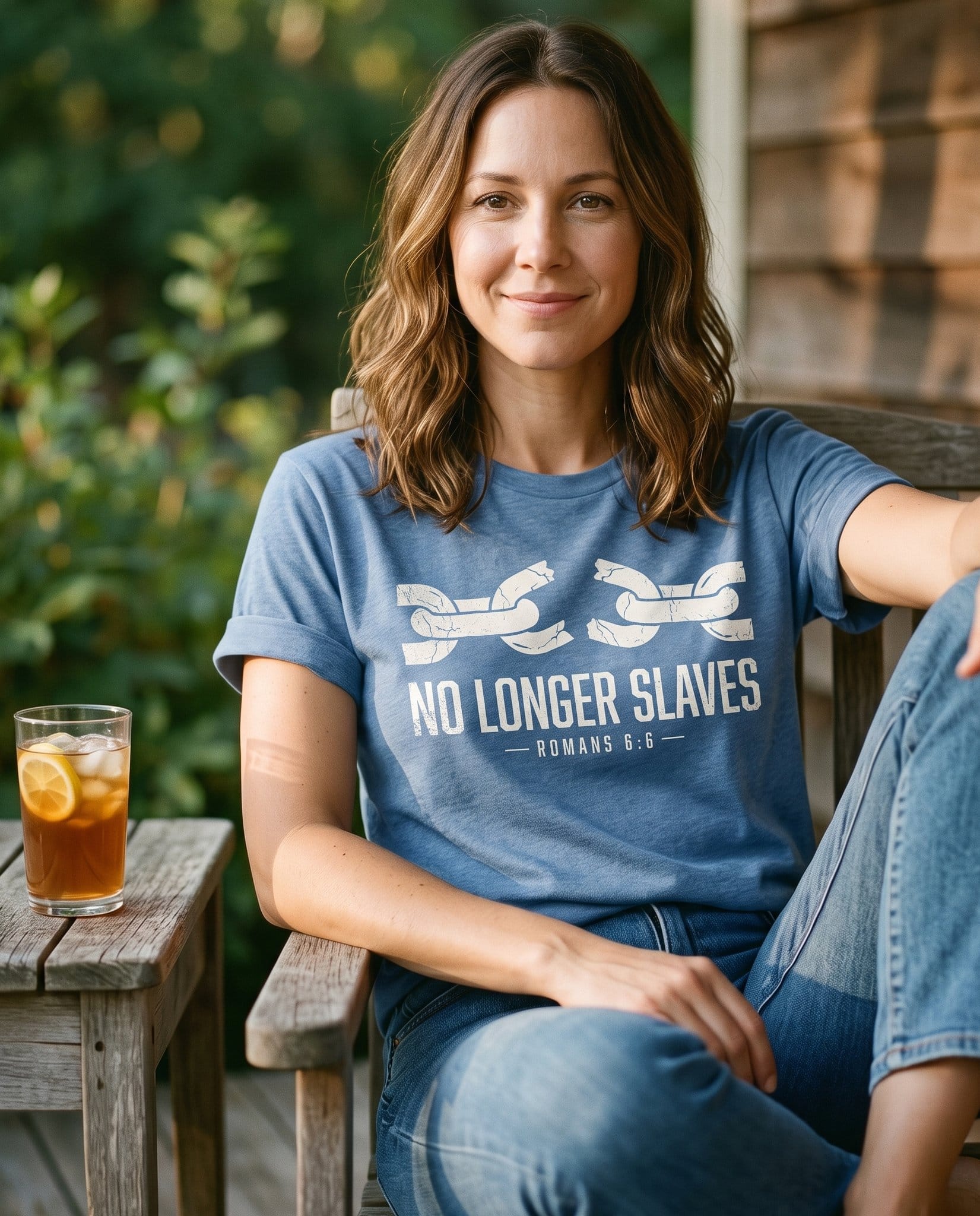No Longer Slaves Romans 6:6 – Blue Jean women's Christian tee, lifestyle photo in sunny porch – A cream illustration of two broken chain links – Romans 6:6 – She Wears Faith