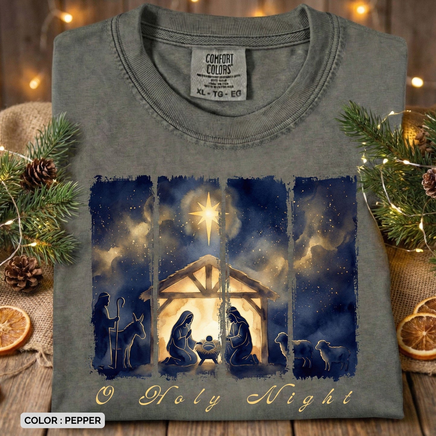 Folded Comfort Colors 1717 pepper Christian women tee showcasing a watercolor “O Holy Night” nativity illustration with Mary, Joseph, baby Jesus, shepherds, and night sky. Cozy and elegant US faith wear photographed with Christmas greenery and warm lights.