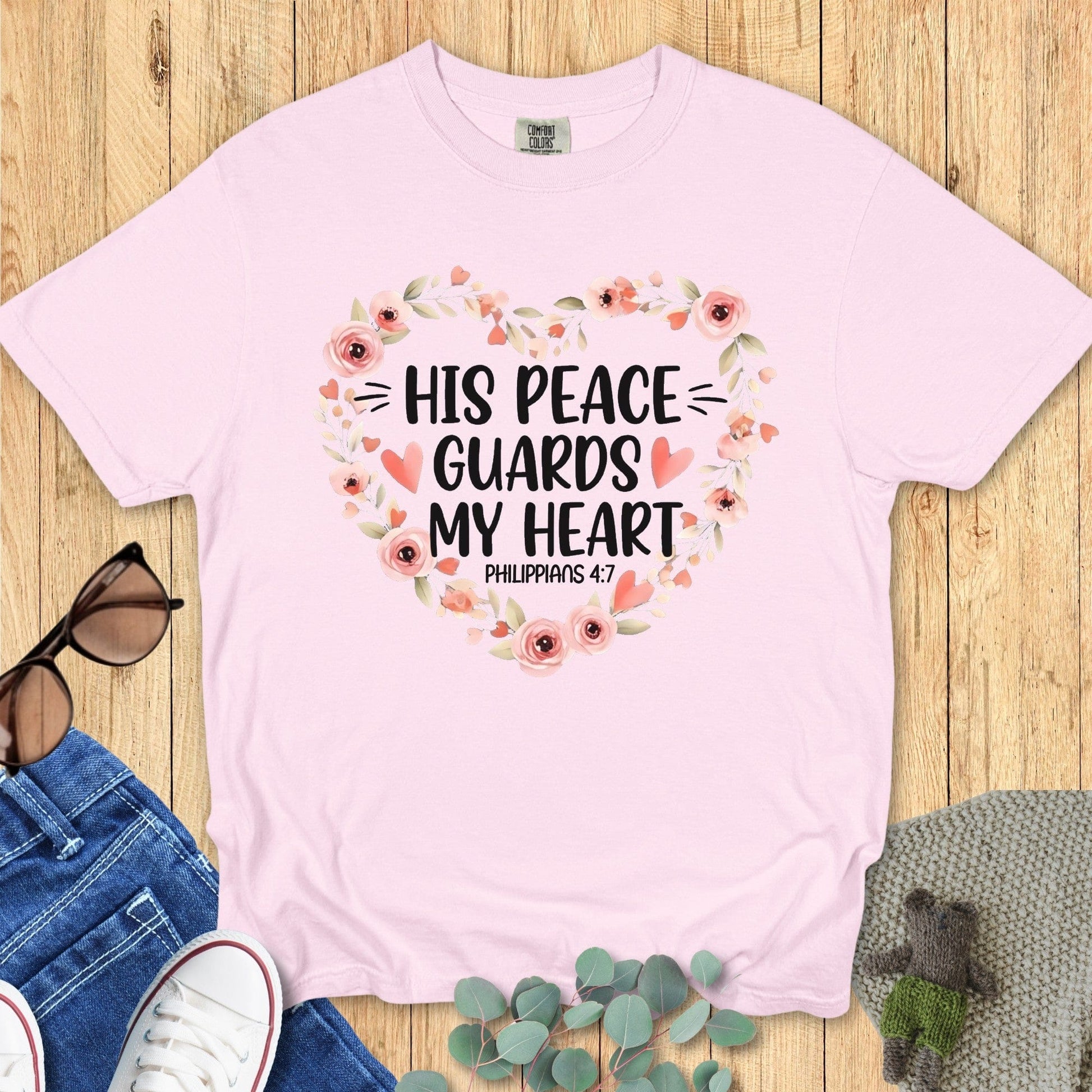 Flatlay of a Comfort Colors 1717 blossom “His Peace Guards My Heart – Philippians 4:7” t-shirt featuring a heart-shaped floral wreath in blush pink tones. A soft and peaceful women’s scripture tee — lovely faith-based apparel for women and a meaningful Christian mom gift idea.