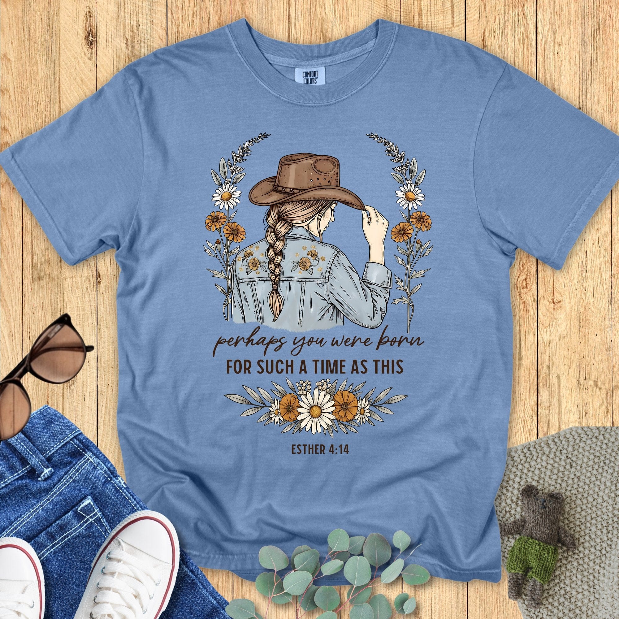 Blue t-shirt with a graphic design of a woman wearing a hat, surrounded by flowers, on a wooden surface.