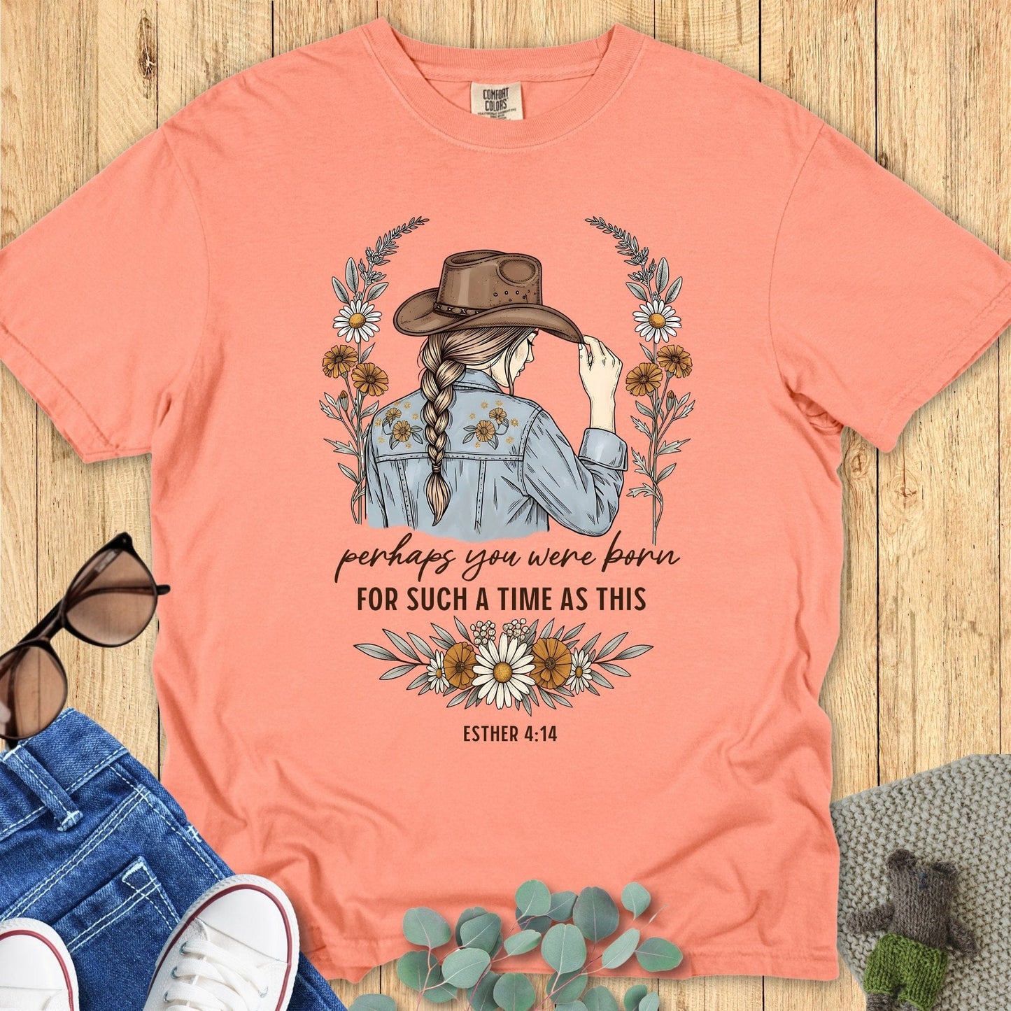 Coral t-shirt with a graphic of a woman wearing a cowboy hat and flowers, text 'perhaps you were born for such a time as this' by Esther 4:14, on a wooden surface with jeans and sunglasses.