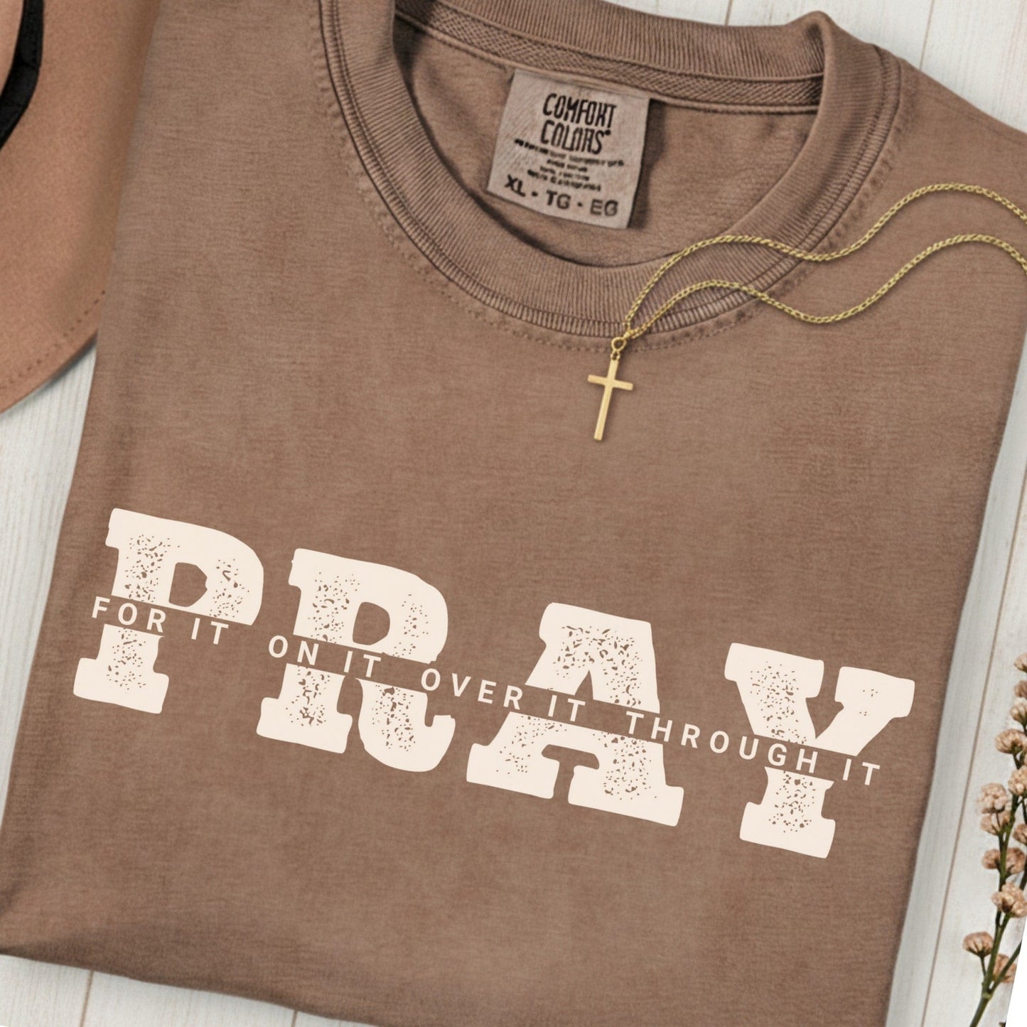 Folded Comfort Colors 1717 espresso “PRAY for it, on it, over it, through it” t-shirt with gold cross necklace on top. A classic women’s scripture tee -  faith-based apparel for women and modern Christian shirt, ideal Christian mom gift idea and prayer reminder.