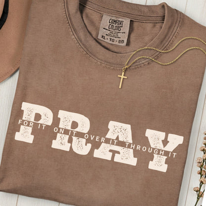 Folded Comfort Colors 1717 espresso “PRAY for it, on it, over it, through it” t-shirt with gold cross necklace on top. A classic women’s scripture tee -  faith-based apparel for women and modern Christian shirt, ideal Christian mom gift idea and prayer reminder.