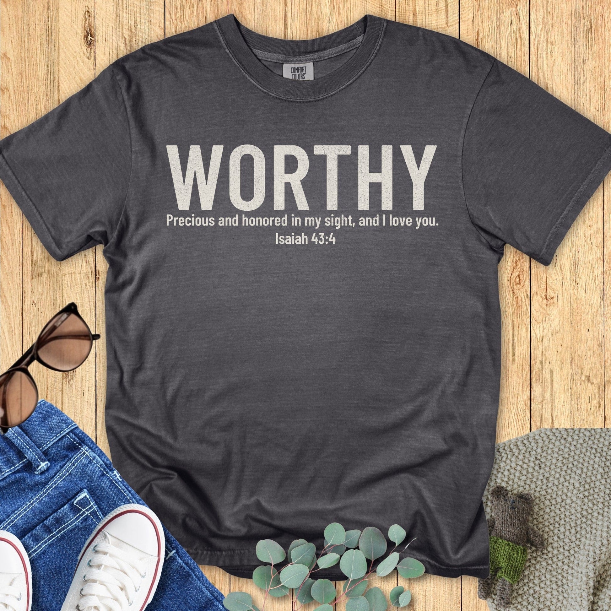Comfort Colors 1717 pepper gray Christian women’s t-shirt reading “WORTHY – Precious and honored in my sight, and I love you. Isaiah 43:4” in distressed white text. Flat lay style with denim, sneakers, sunglasses, and eucalyptus leaves on rustic wood