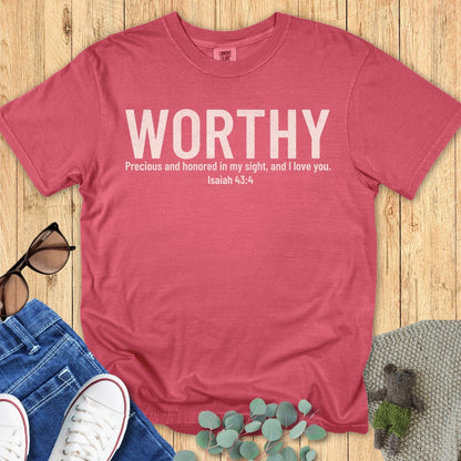 Christian women’s Comfort Colors 1717 crimson red t-shirt with “WORTHY” in large white distressed letters, and Isaiah 43:4 verse beneath: “Precious and honored in my sight, and I love you.” Styled flat with casual accessories on wooden backdrop