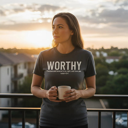 Woman standing on a balcony at sunset wearing a Comfort Colors 1717 charcoal Christian t-shirt with bold white text that reads “WORTHY – Precious and honored in my sight, and I love you. Isaiah 43:4.” She holds a coffee mug, city skyline and golden sky in background