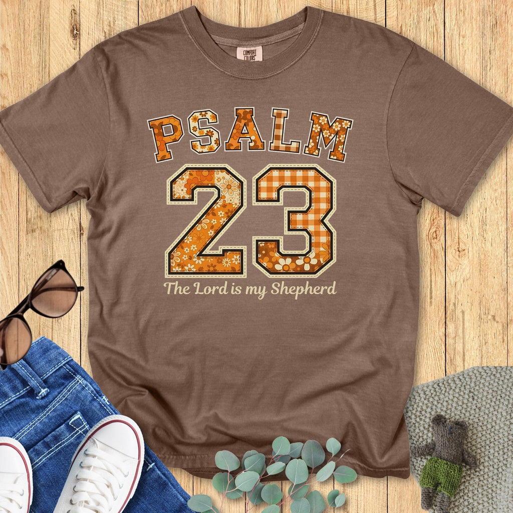 Flatlay of a Comfort Colors 1717 expresso “Psalm 23 — The Lord is my Shepherd” t-shirt featuring floral and gingham varsity lettering in warm tones. A classic women’s scripture tee - faith-based apparel for women and modern Christian shirt, lovely Christian mom gift idea for fall or church events.