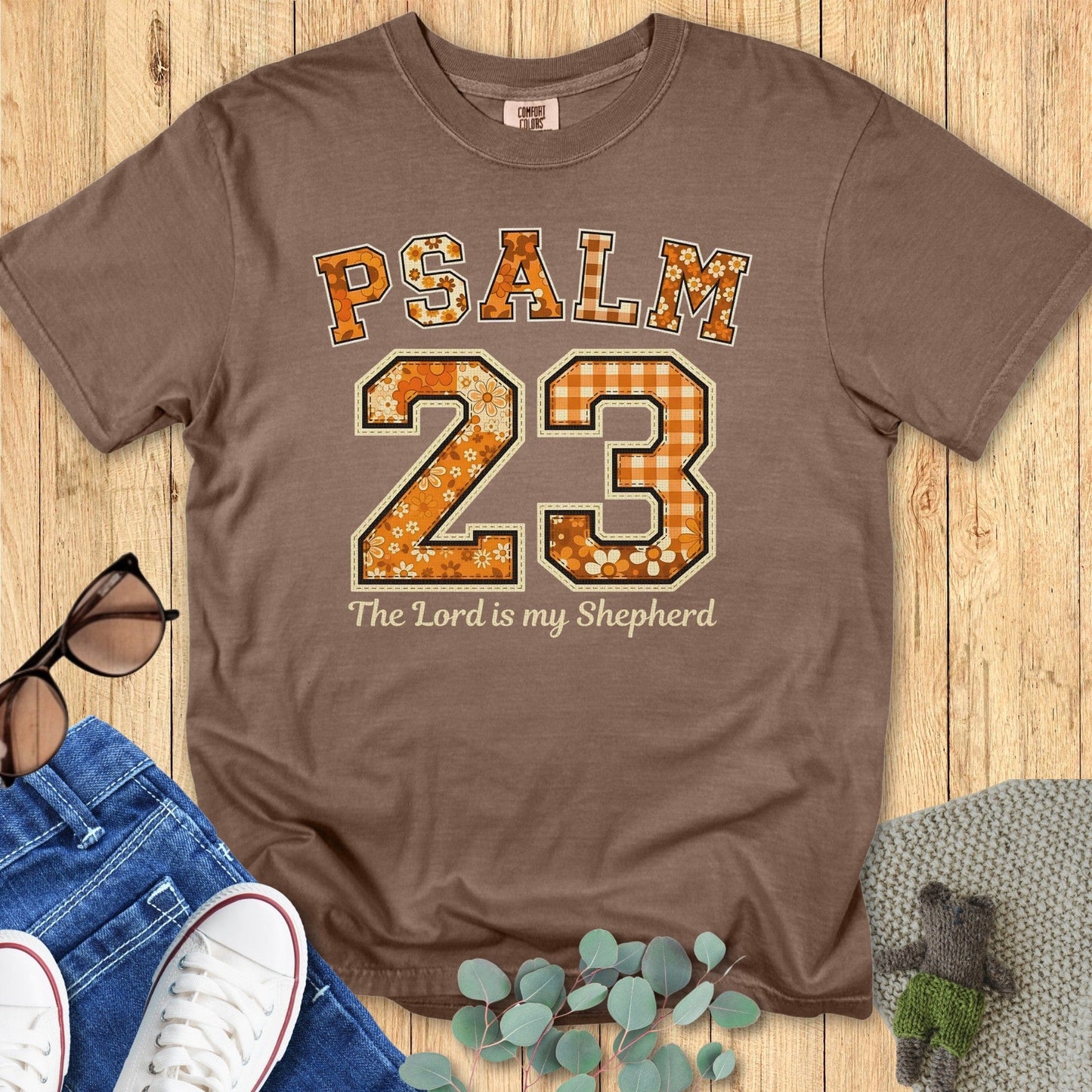 Flatlay of a Comfort Colors 1717 expresso “Psalm 23 — The Lord is my Shepherd” t-shirt featuring floral and gingham varsity lettering in warm tones. A classic women’s scripture tee - faith-based apparel for women and modern Christian shirt, lovely Christian mom gift idea for fall or church events.