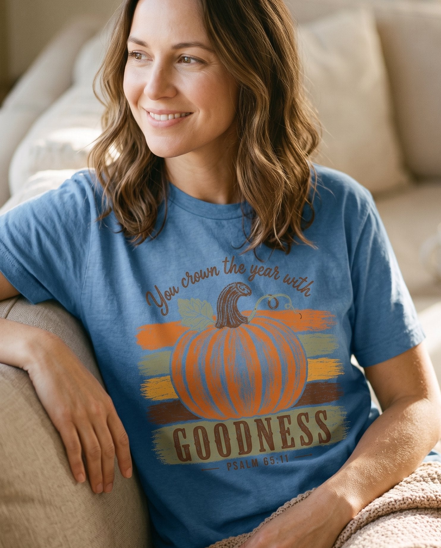 Pumpkin Goodness Psalm 65 11 Goodness – Blue Jean women's Christian tee, lifestyle photo in cozy living room – Hand-sketched orange pumpkin against layered autumn brushstroke stripe – Psalm 65:11 – She Wears Faith