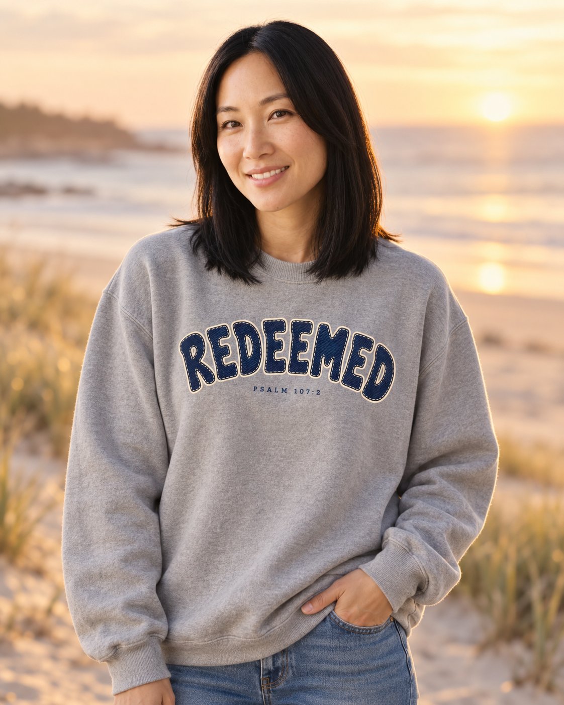 Redeemed Psalm 107:2, Grey women's Christian sweatshirt, lifestyle photo in seaside setting, Collegiate stitched REDEEMED lettering, She Wears Faith
