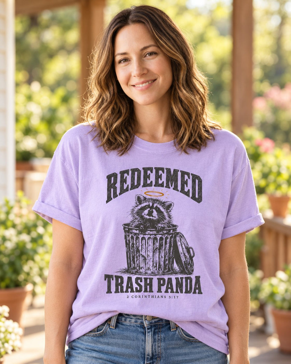 Redeemed Trash Panda, Orchid women's Christian tee, lifestyle photo in sunny porch, haloed raccoon in a trash can with REDEEMED TRASH PANDA, 2 Corinthians 5:17, She Wears Faith