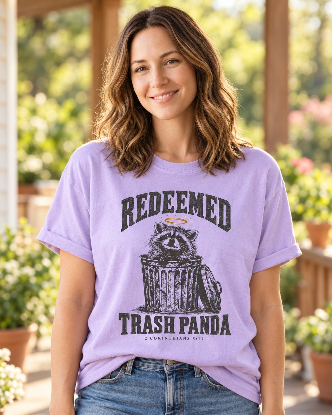 Redeemed Trash Panda, Orchid women's Christian tee, lifestyle photo in sunny porch, haloed raccoon in a trash can with REDEEMED TRASH PANDA, 2 Corinthians 5:17, She Wears Faith