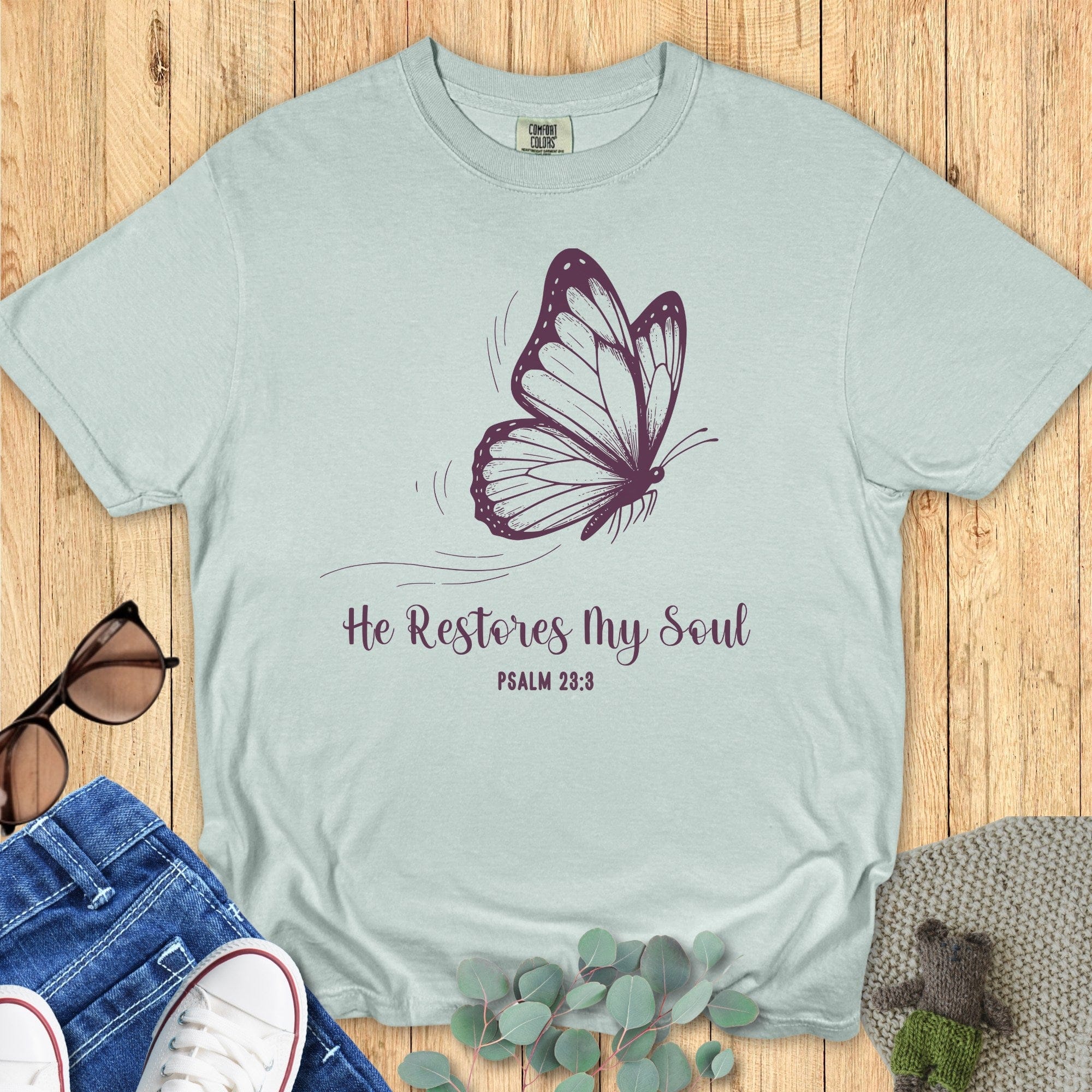 Flatlay of a Comfort Colors 1717 bay “He Restores My Soul – Psalm 23:3” t-shirt in soft mint tone with graceful butterfly print. A calming women’s scripture tee — modern faith-based apparel for women and meaningful Christian mom gift idea inspired by Psalm 23:3.