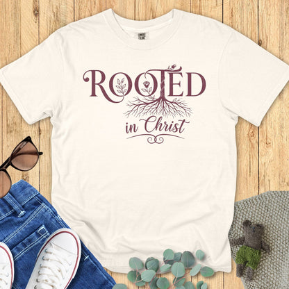 “Ivory Comfort Colors 1717 Christian T-shirt with ‘Rooted in Christ’ design in maroon lettering and tree roots graphic, styled with denim jeans and sneakers on a wooden background.”
