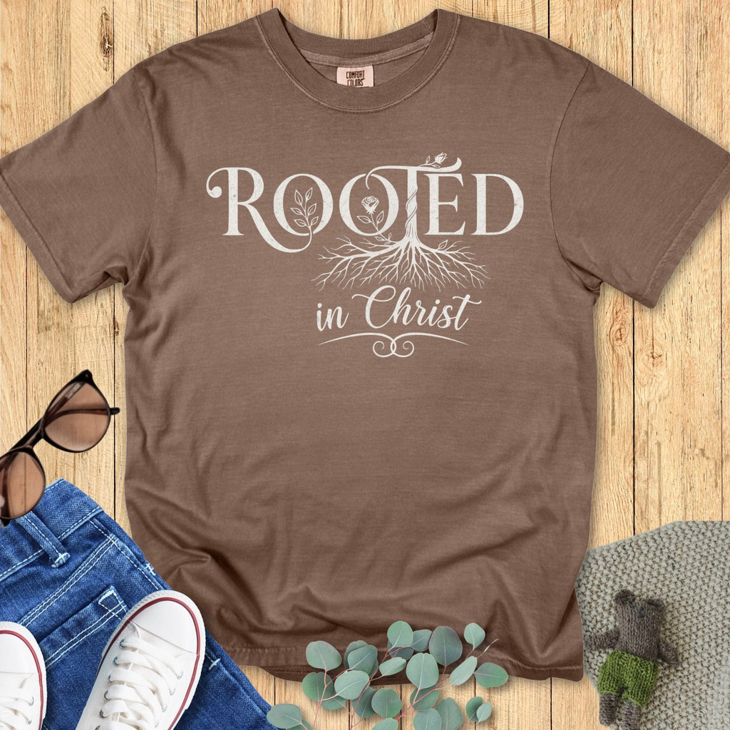 Espresso brown Comfort Colors 1717 Christian T-shirt with ‘Rooted in Christ’ lettering and roots graphic, styled in a flat lay with sunglasses, jeans, and greenery accents.”