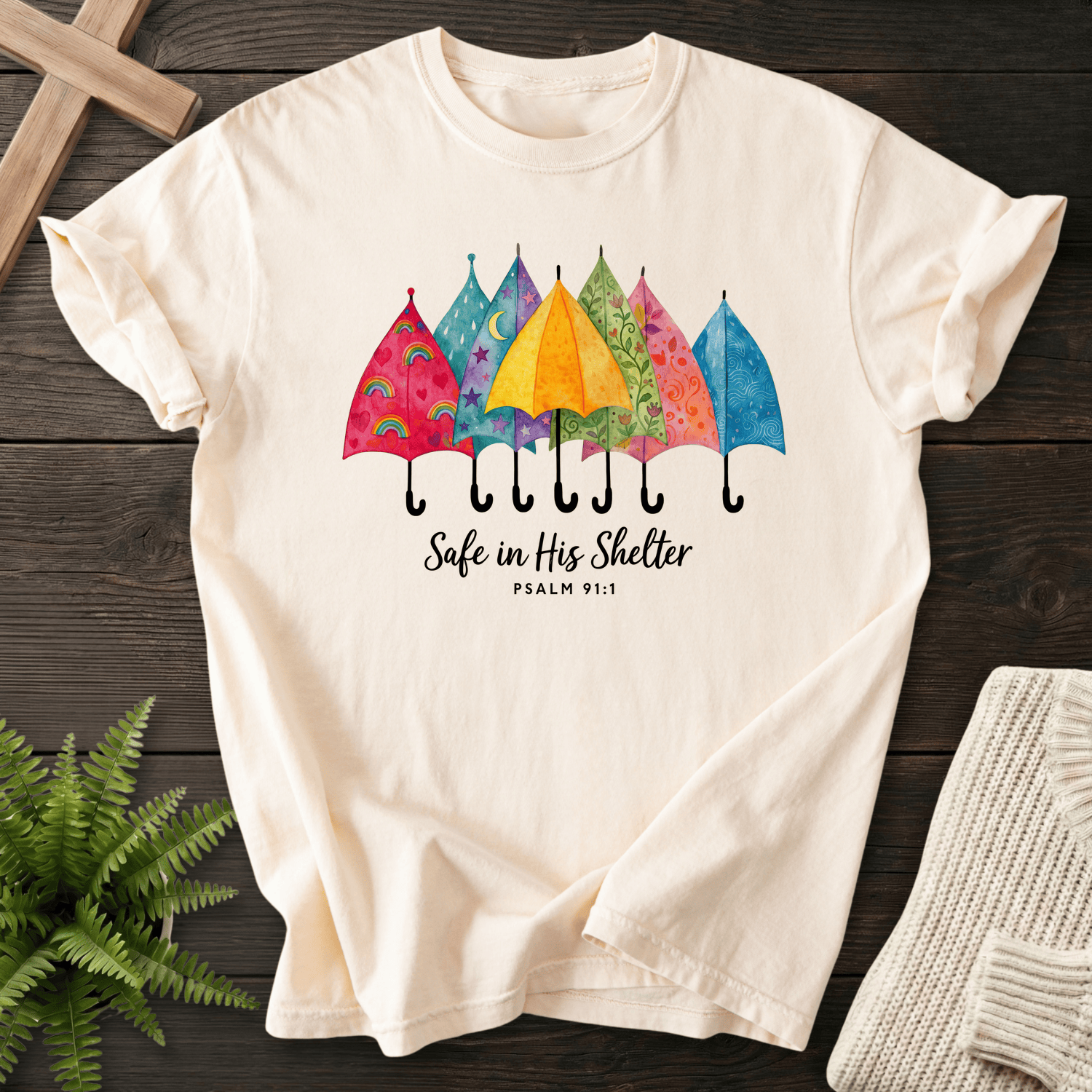 Ivory women's Christian t-shirt with watercolor umbrella design and "Safe in His Shelter," inspired by Psalm 91:1.