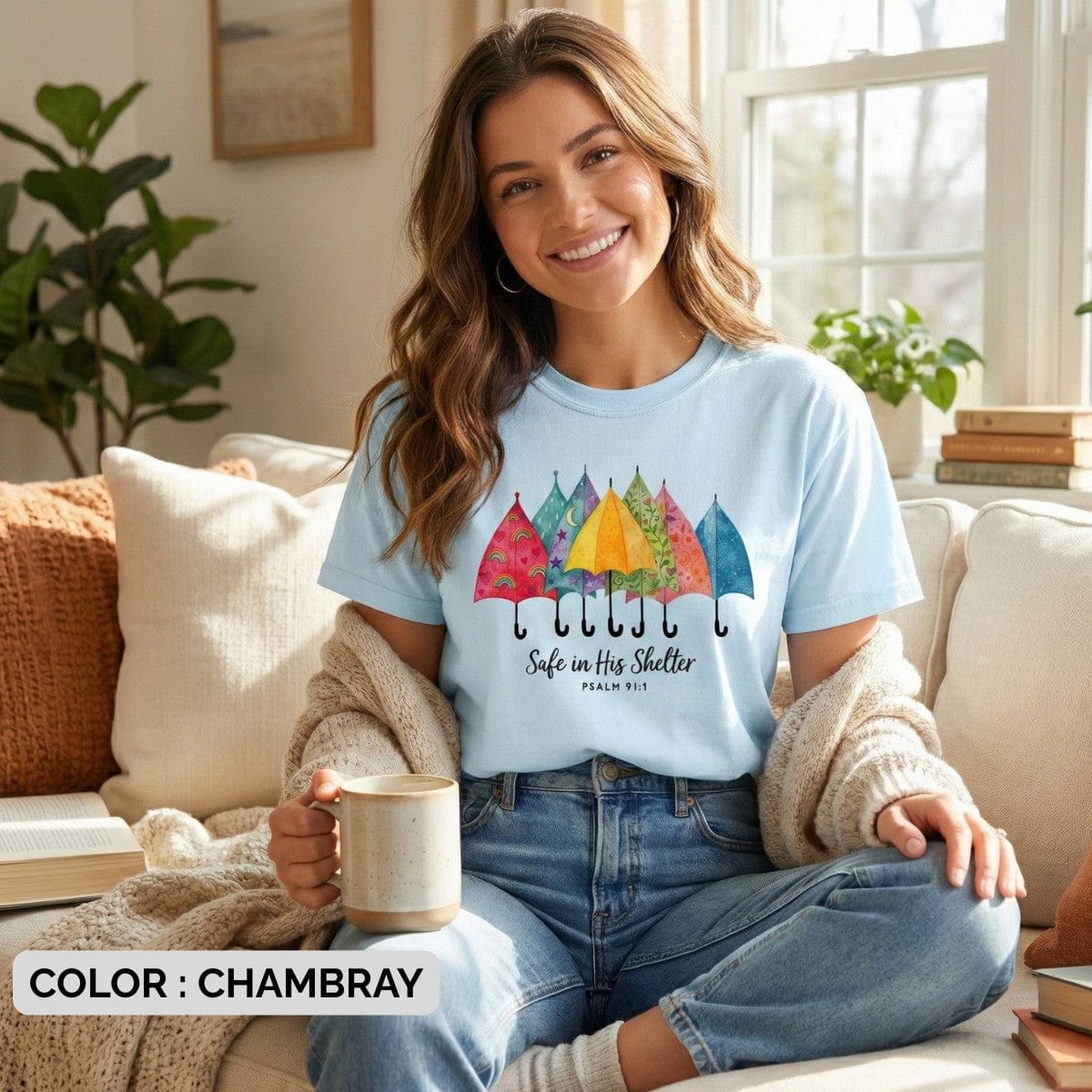Smiling woman sitting on a sofa wearing a Chambray Comfort Colors 1717 tshirt. Lifestyle photography of a Christian women tee featuring "Safe in His Shelter," showcasing casual US faith wear for the home.