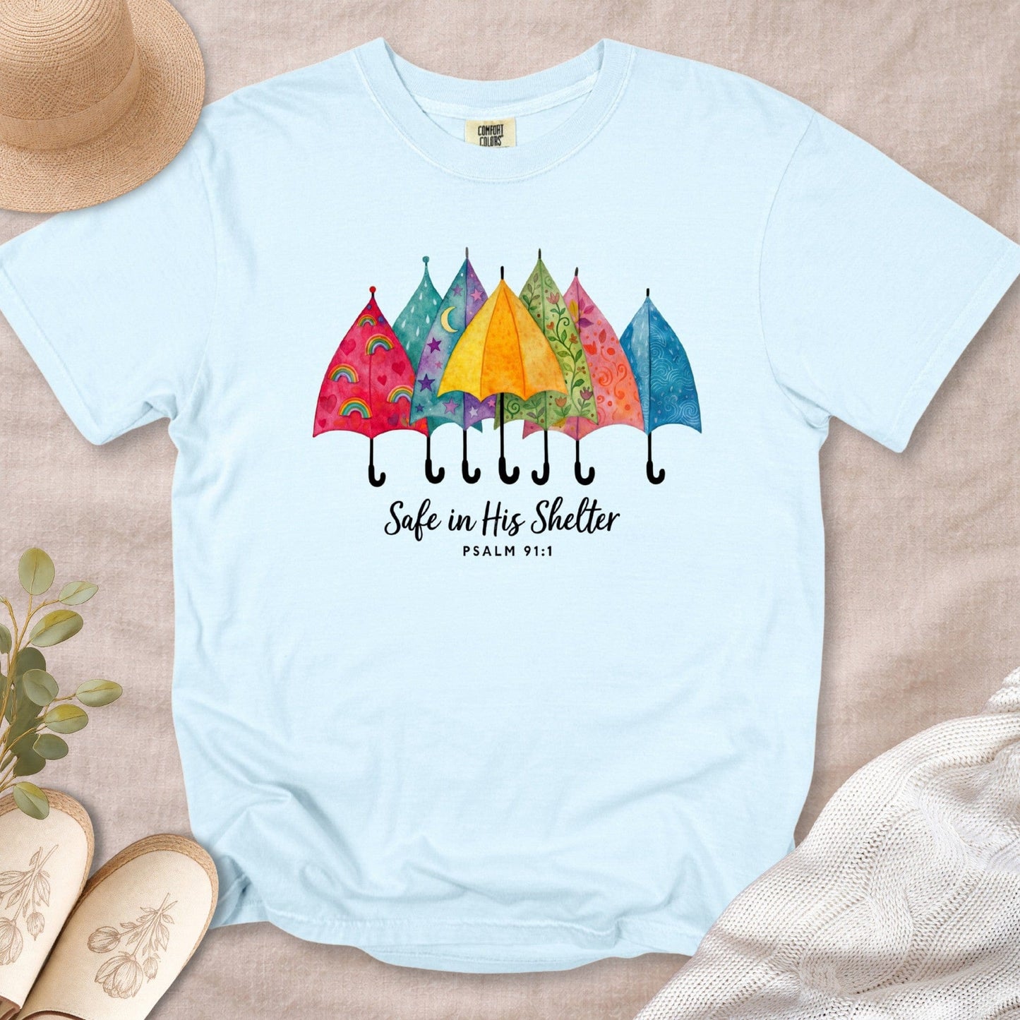 Chambray Blue Comfort Colors 1717 tshirt displaying a whimsical umbrella illustration and Bible verse. Soft Christian women tee with the text "Safe in His Shelter," styled for US faith wear shops.