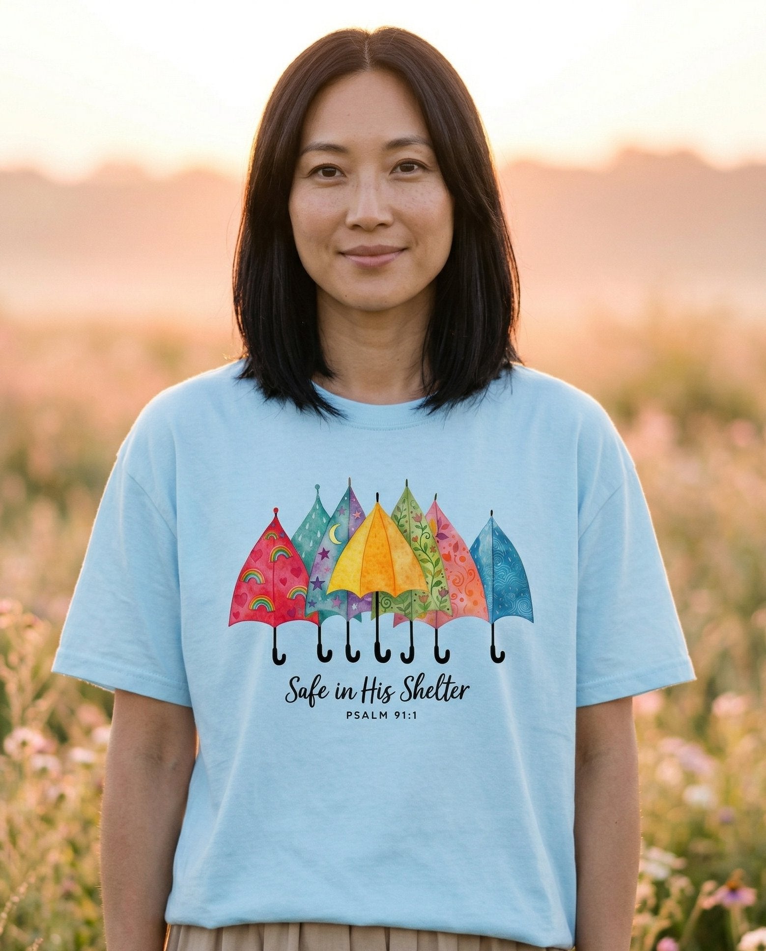 Safe in His Shelter – Chambray women's Christian tee, lifestyle photo in wildflower meadow at sunrise – Colorful watercolor row of six patterned umbrellas in reds – Psalm 91:1 – She Wears Faith