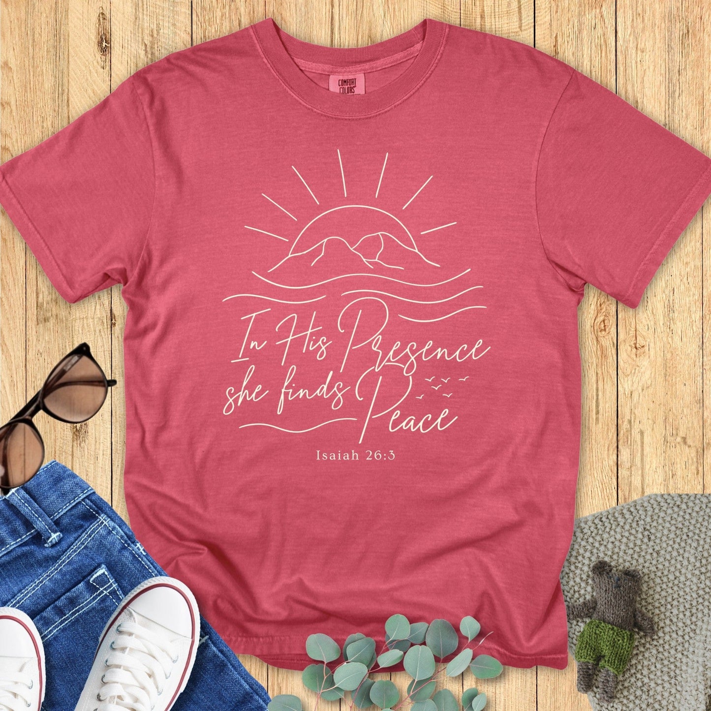 Flatlay of a Comfort Colors 1717 crimson “In His Presence She Finds Peace – Isaiah 26:3” t-shirt with simple sun and ocean waves design. A peaceful and encouraging women’s scripture tee — modern Christian shirt and beautiful faith-based apparel for women.