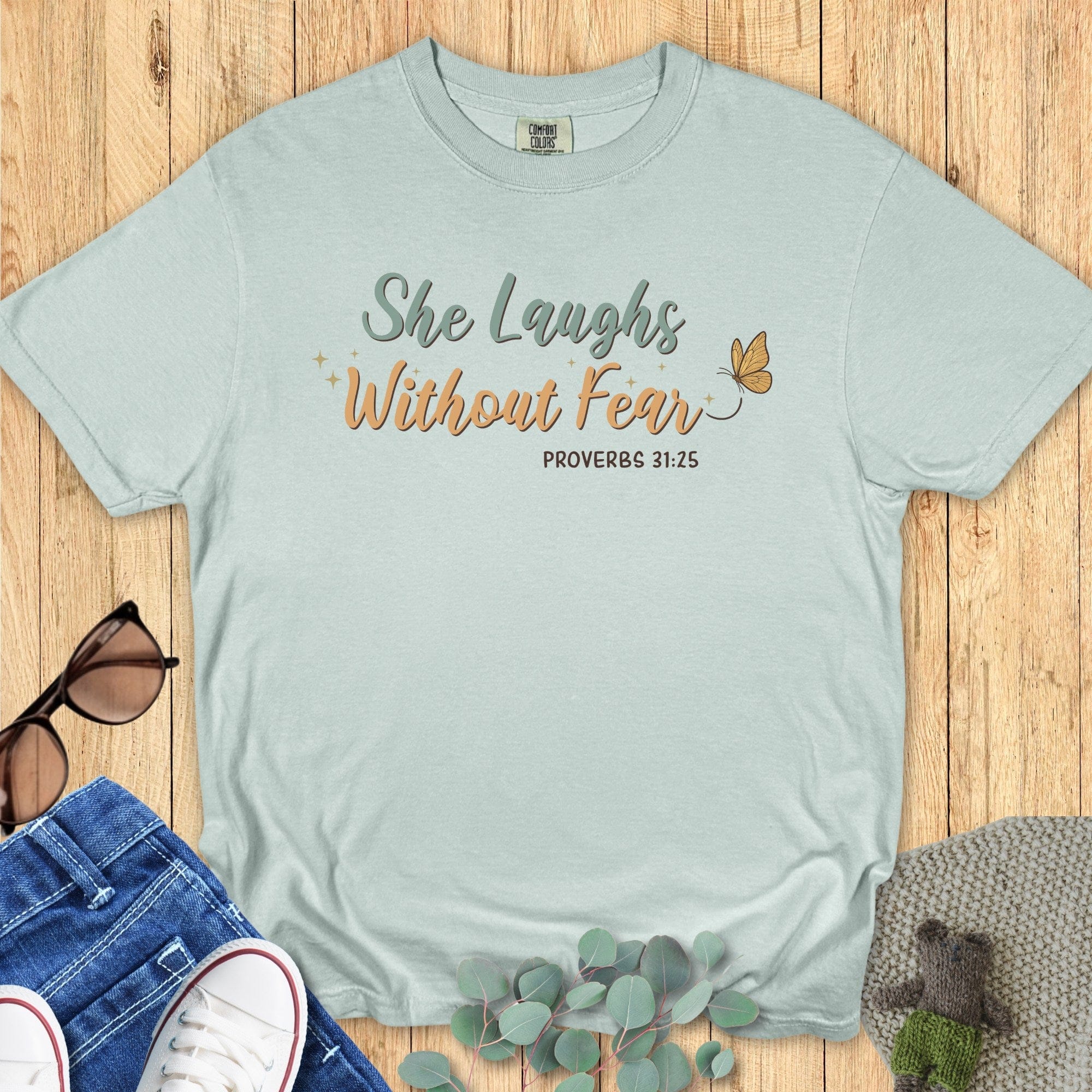 Flatlay of a Comfort Colors 1717 bay “She Laughs Without Fear – Proverbs 31:25” t-shirt featuring soft script typography and a butterfly accent. A stylish women’s scripture tee — faith-based apparel for women and a thoughtful Christian mom gift idea.