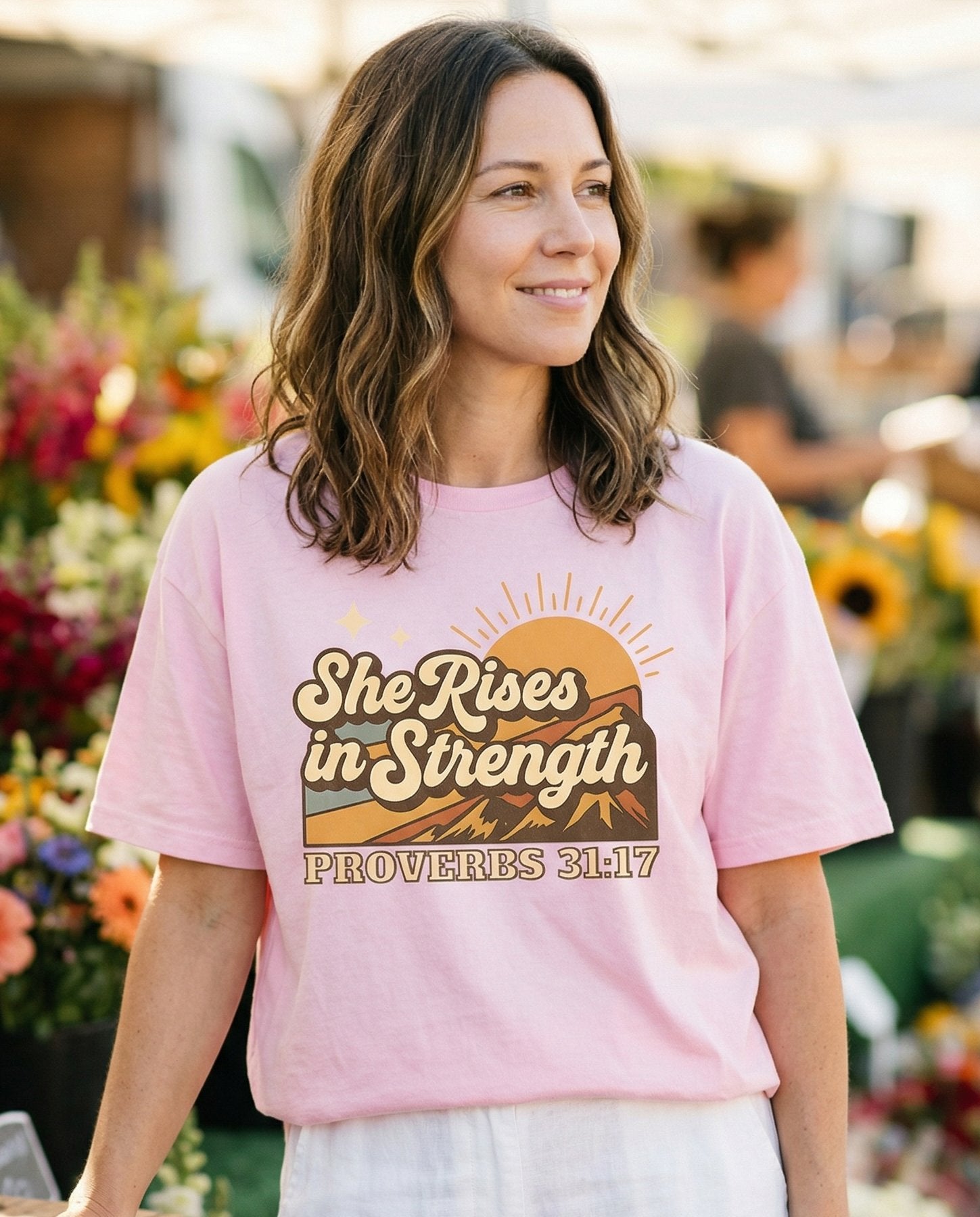 She Rises in Strength – Blossom women's Christian tee, lifestyle photo in farmers market – Retro vintage-style graphic with mountains – Proverbs 31:17 – She Wears Faith