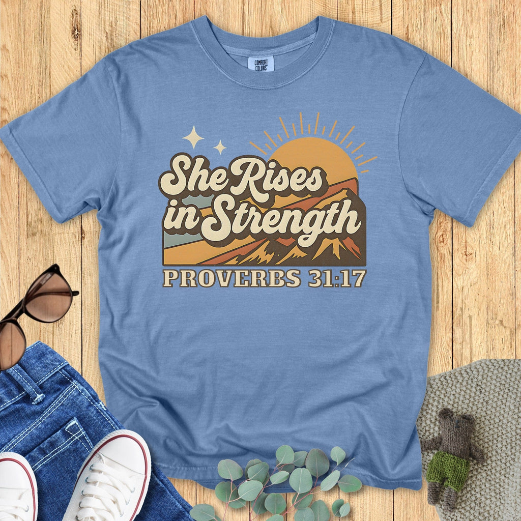 Flatlay of a Comfort Colors 1717 blue jean “She Rises in Strength – Proverbs 31:17” t-shirt with warm-toned mountain sunrise design. A faith-filled women’s scripture tee — durable faith-based apparel for women and encouraging modern Christian shirt, great for Christian mom gift ideas.
