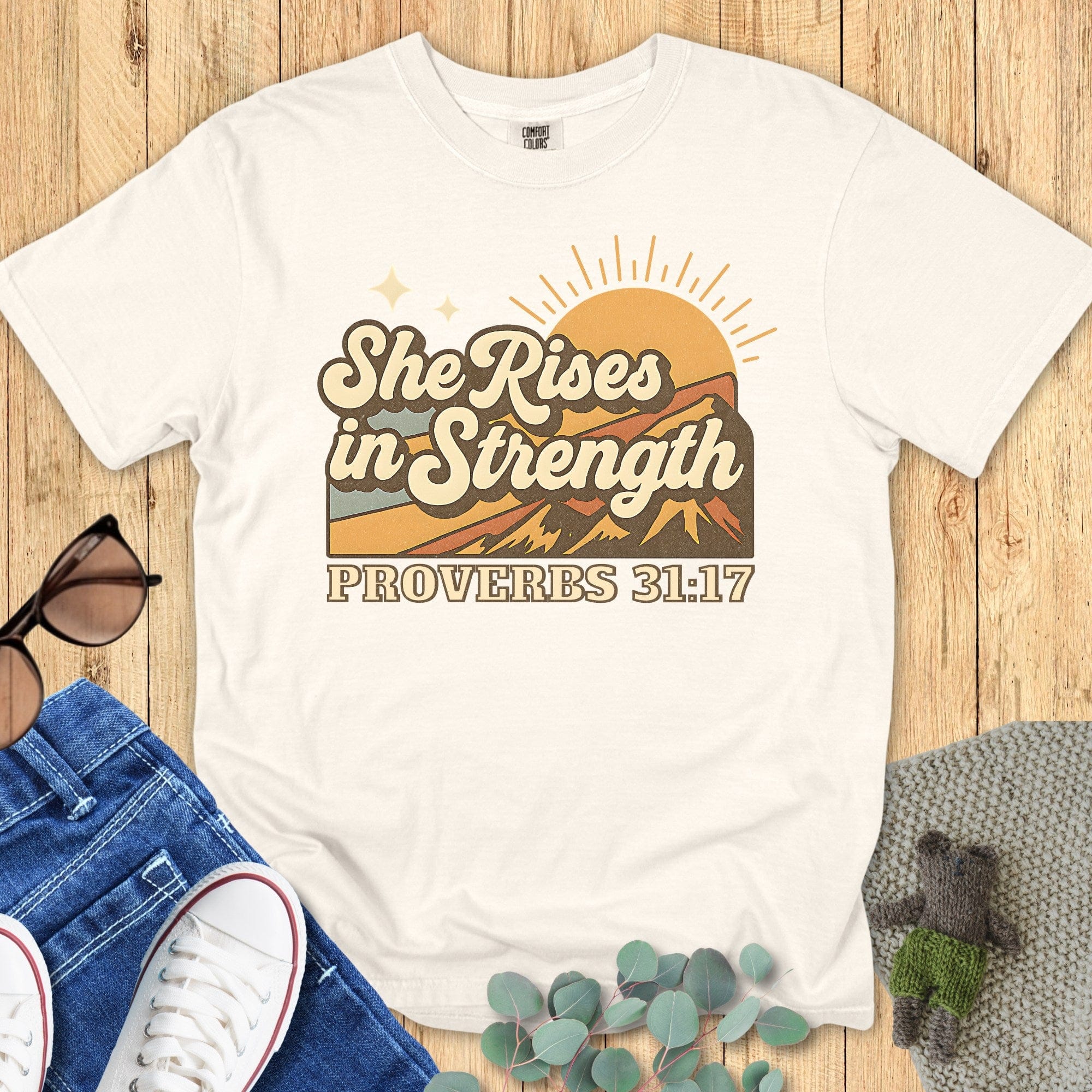 Flatlay of a Comfort Colors 1717 ivory “She Rises in Strength – Proverbs 31:17” t-shirt featuring a vintage mountain sunrise graphic. A stylish women’s scripture tee — soft faith-based apparel for women and uplifting modern Christian shirt, ideal Christian mom gift idea.