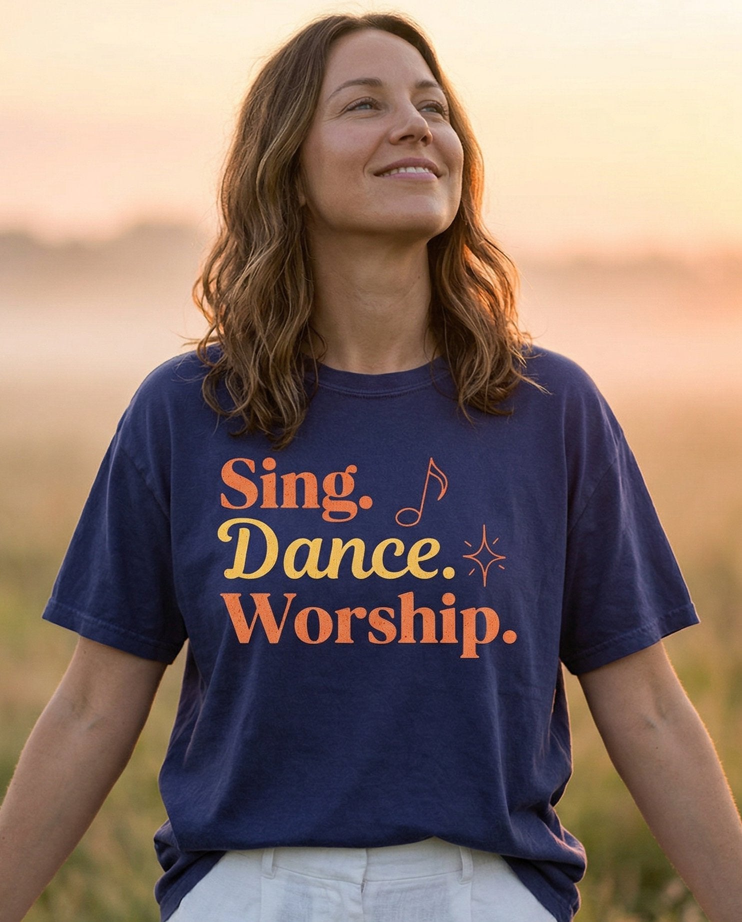 Sing, Dance, Worship T-shirt – Grape women's Christian tee, lifestyle photo in wildflower meadow at sunrise – Bold retro-style stacked lettering 'Sing – She Wears Faith